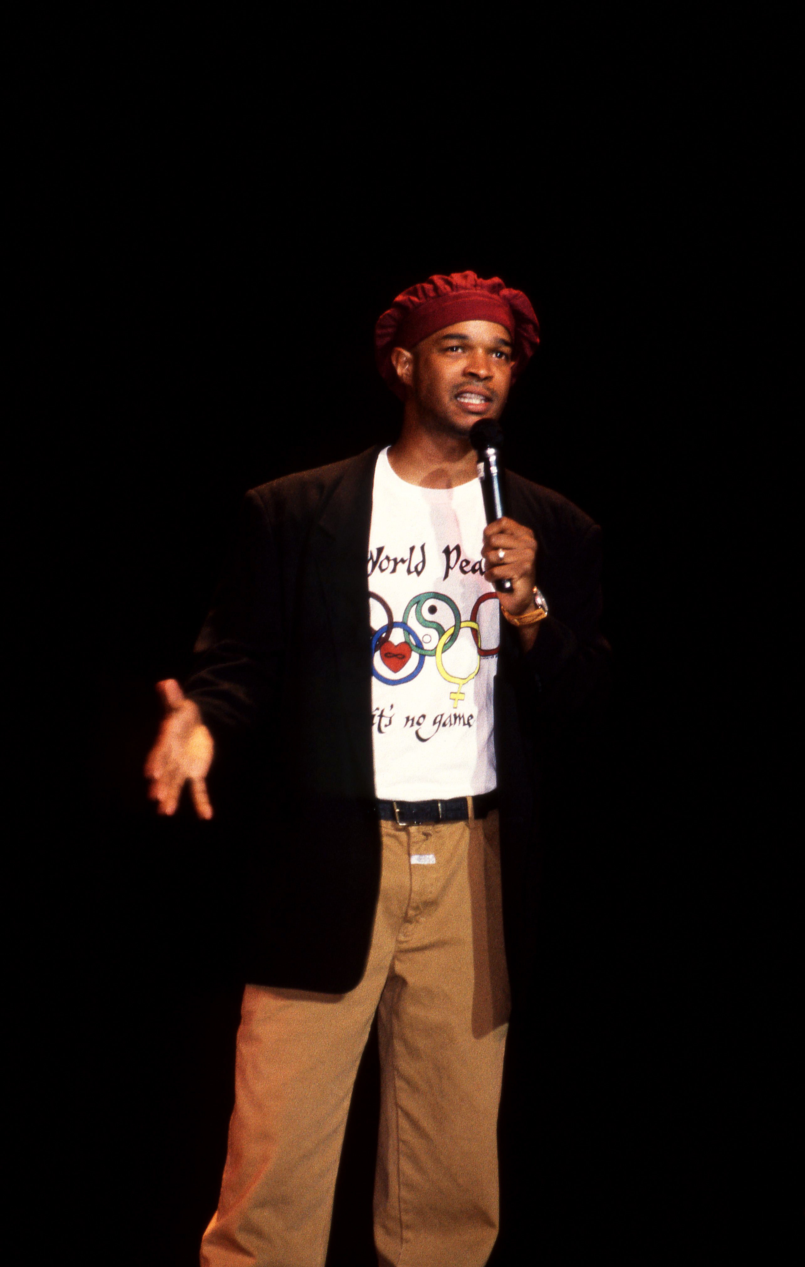 Person on stage wearing a beret, printed t-shirt with peace symbols, blazer, and pants, holding a microphone, performing or speaking