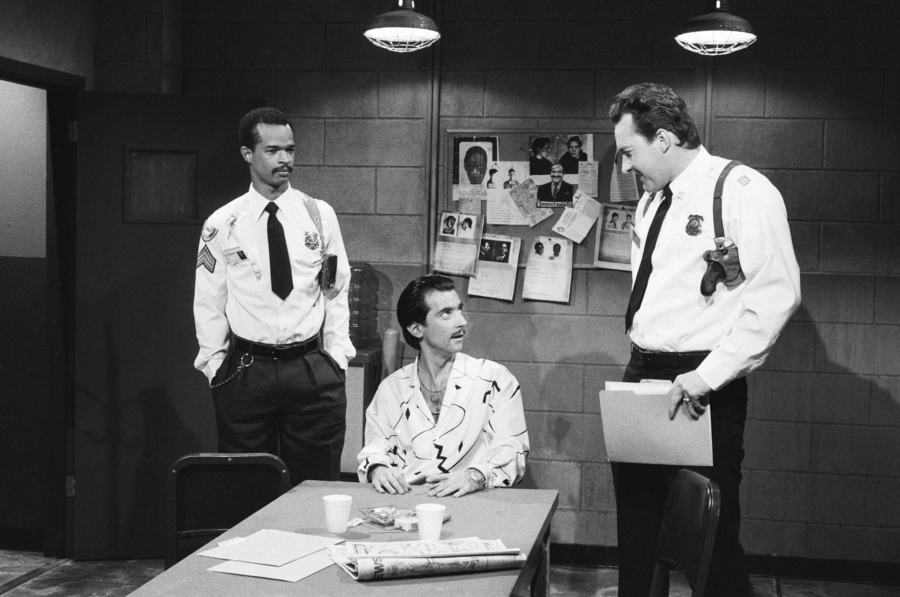 Three men in police uniforms are in an office, engaged in conversation around a table with papers and a coffee cup. Bulletin board in the background