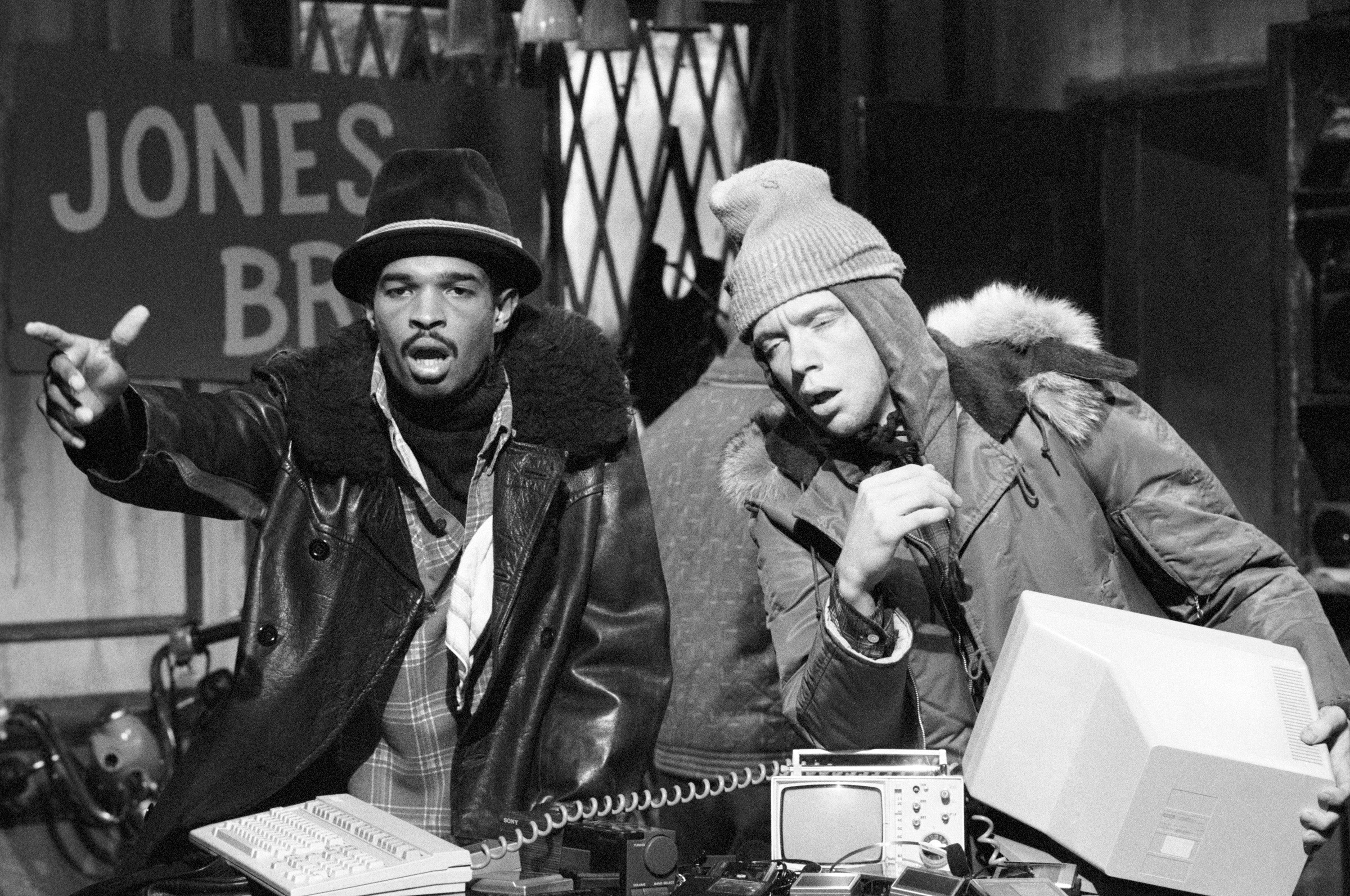 Two people on a TV set resembling a pawn shop, wearing winter coats and hats, looking animated while gesturing and talking