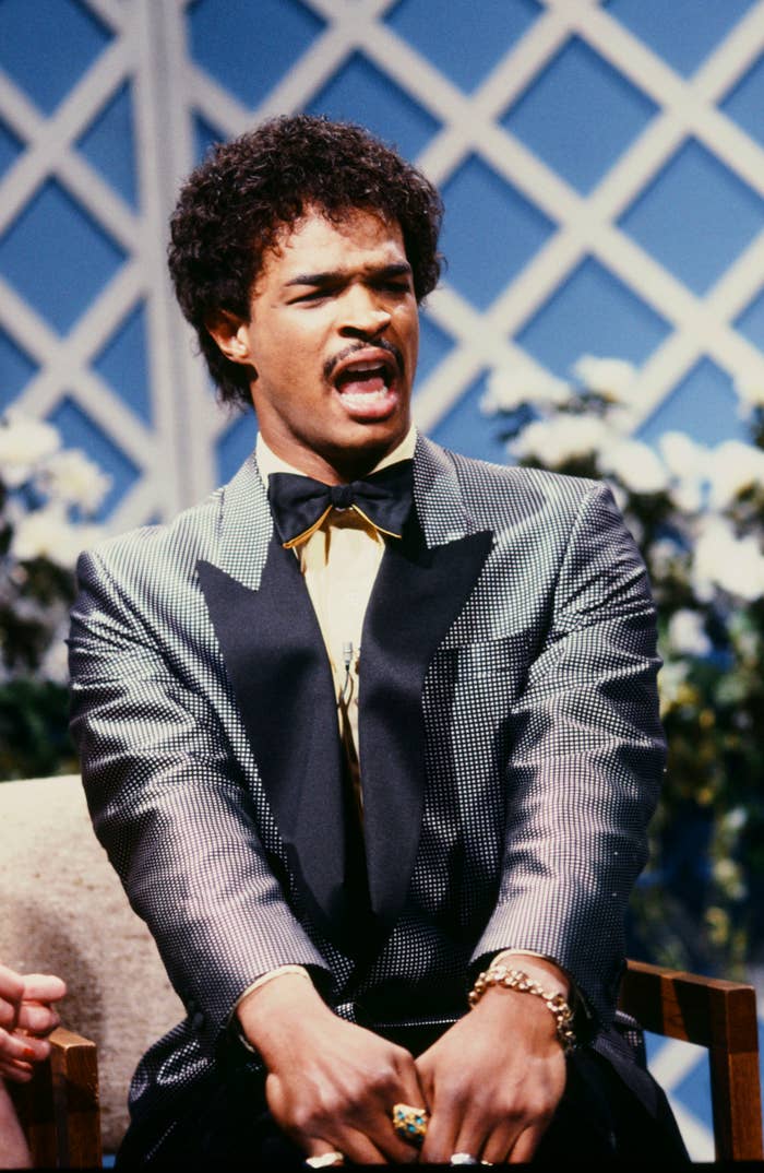 A person in a shiny tuxedo with wide lapels and a bow tie, sitting and gesturing expressively on a set with a lattice backdrop