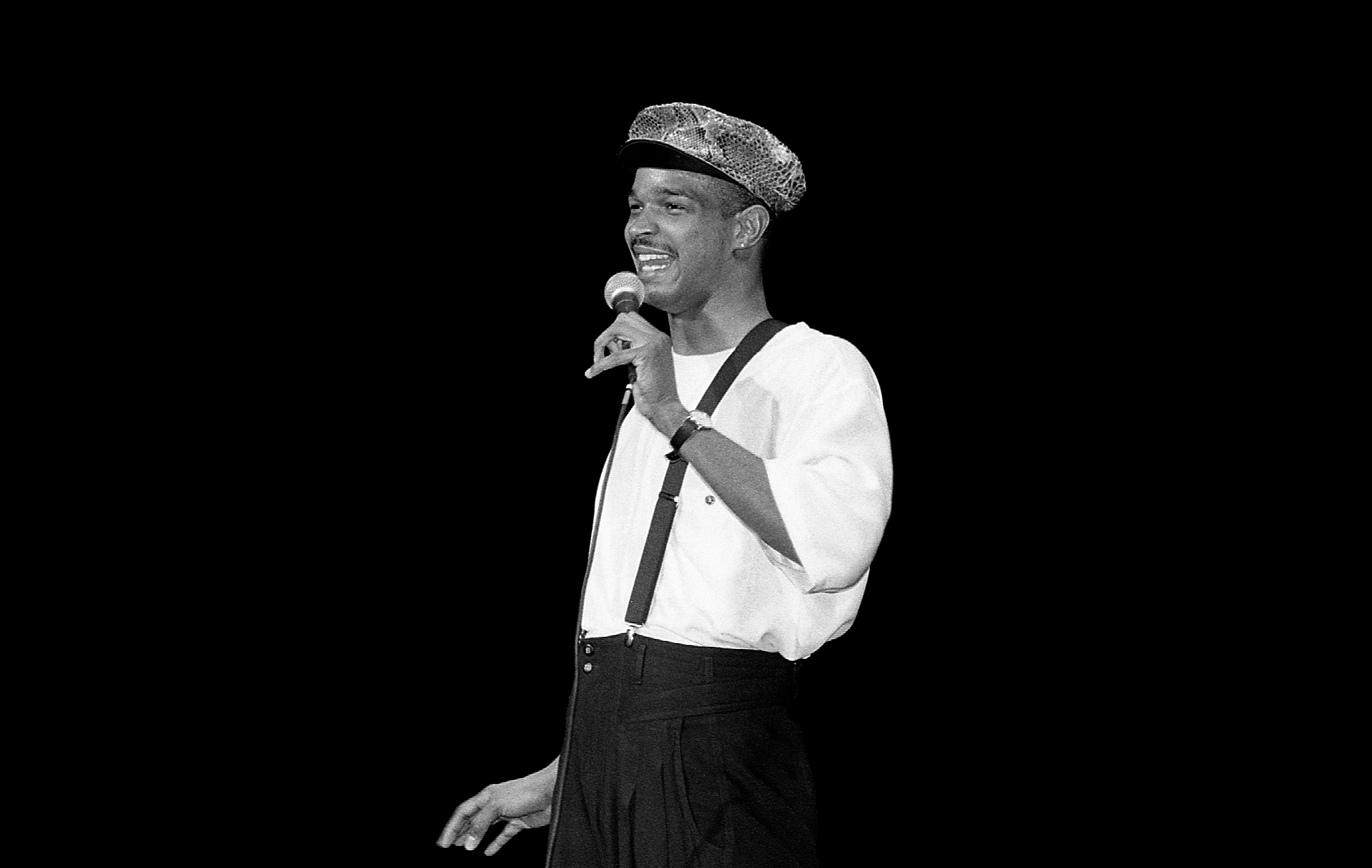 Person on stage in vintage-inspired outfit, wearing suspenders and a cap, holding a microphone, smiling as they perform