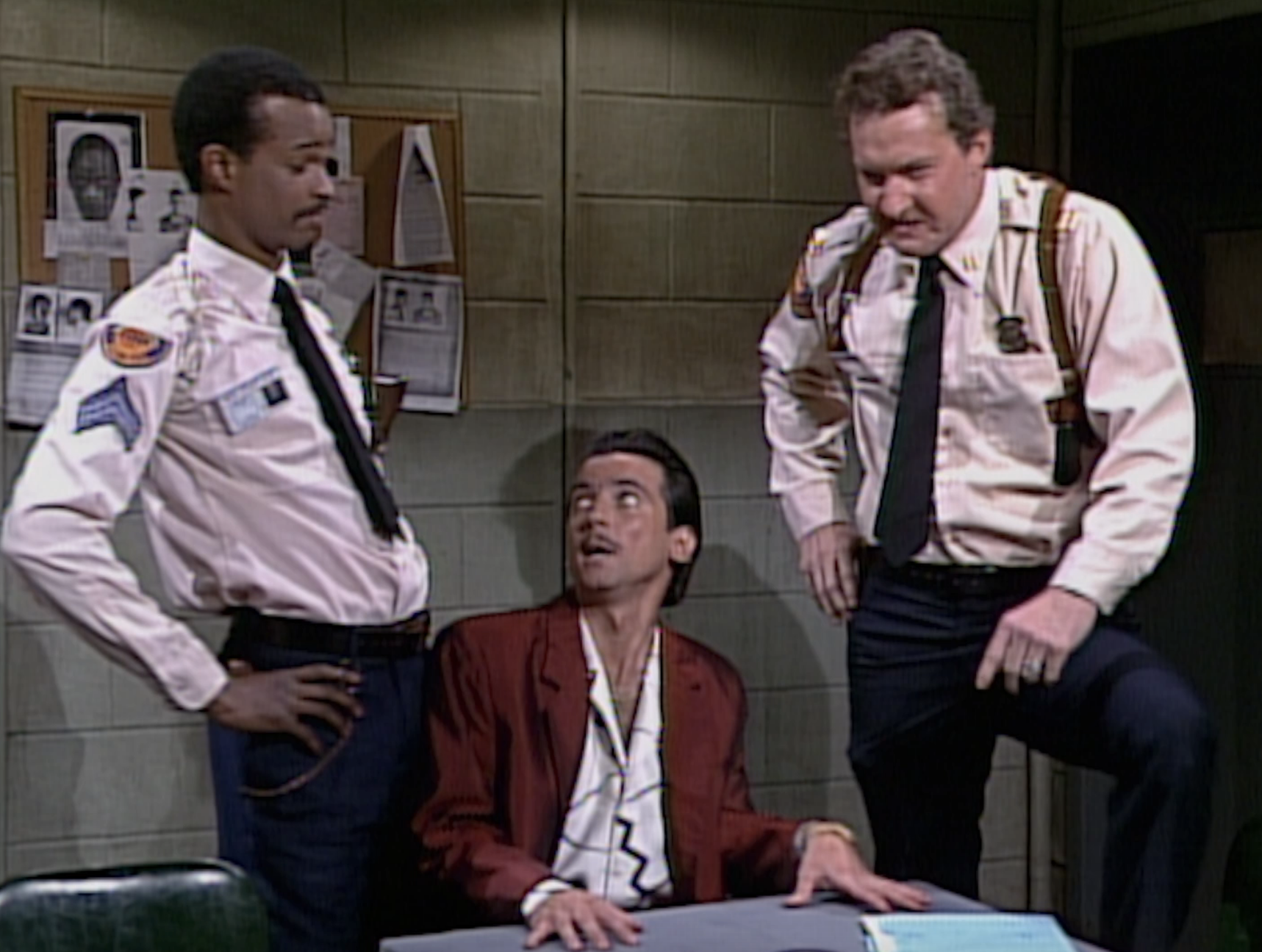 Three men in a TV crime scene; one seated in a red jacket, two standing in police uniforms in what appears to be an interrogation room