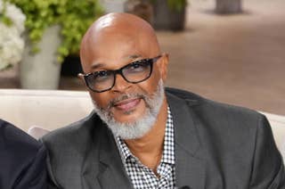Man with glasses and gray beard smiles, wearing a checked shirt and dark blazer, sitting in a cozy setting