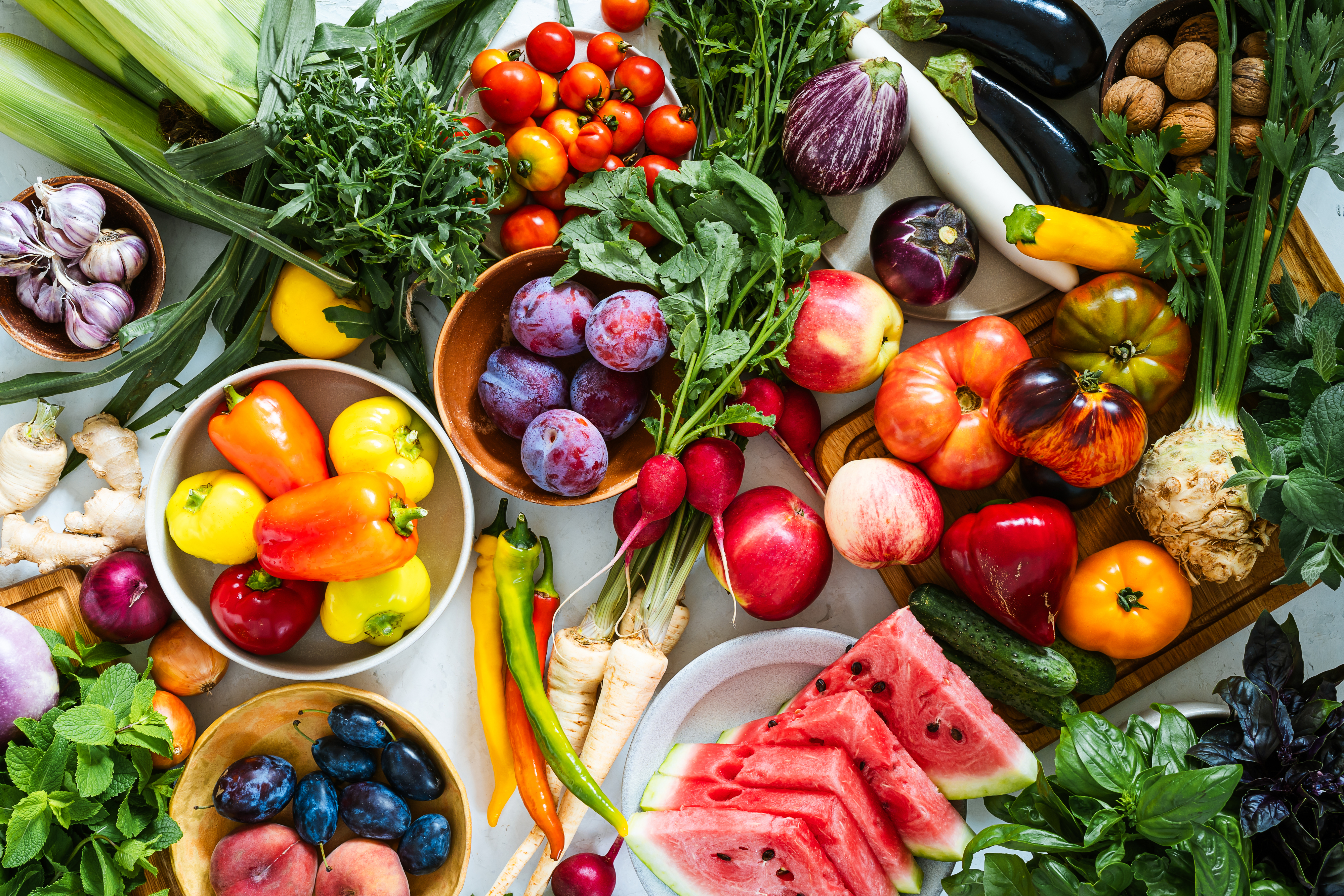 Assorted fresh produce including tomatoes, plums, watermelon, peppers, radishes, and herbs displayed on a table, highlighting seasonal abundance