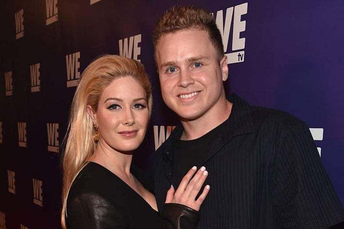 Two people pose together at a WE TV event. One is wearing a black outfit with leather accents; the other is in a dark button-up shirt