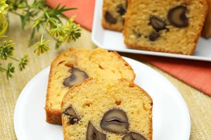 ケーキのスライスが皿に乗っており、ナッツが生地に入っている。隣に植物が飾られている。