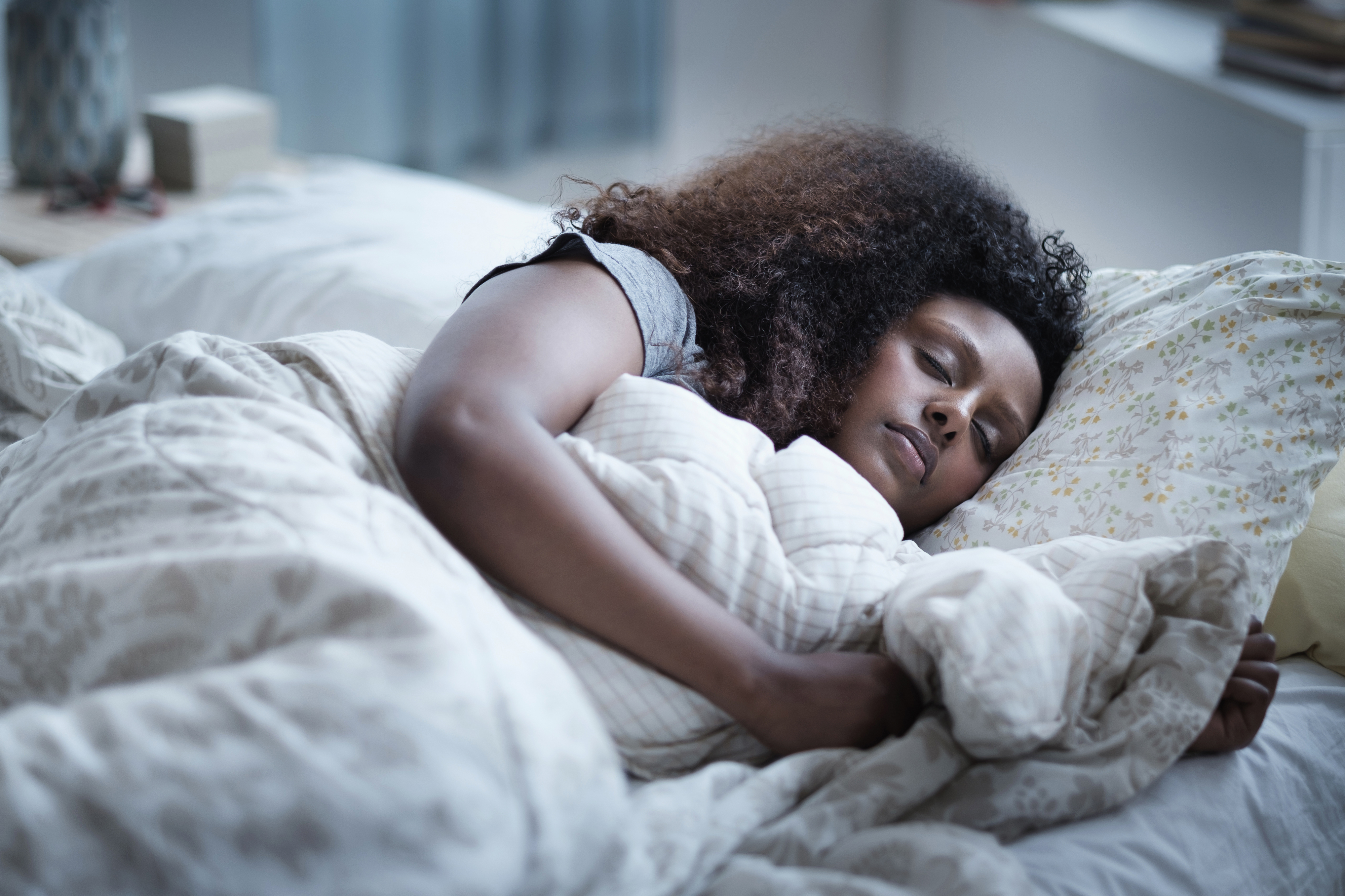 Person som sover fredelig i en seng, omfavnet av mønstrede tepper, med naturlig lysfiltrering gjennom et nærliggende vindu