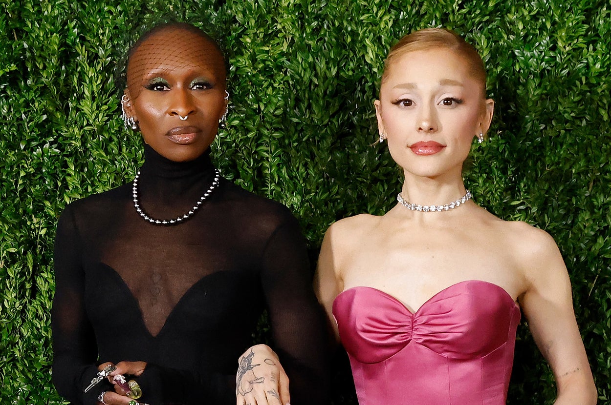 Ariana Grande and Cynthia Erivo in elegant dresses on a lush green backdrop: Cynthia in black with a sheer top, Ariana in a strapless pink gown, both with jewelry