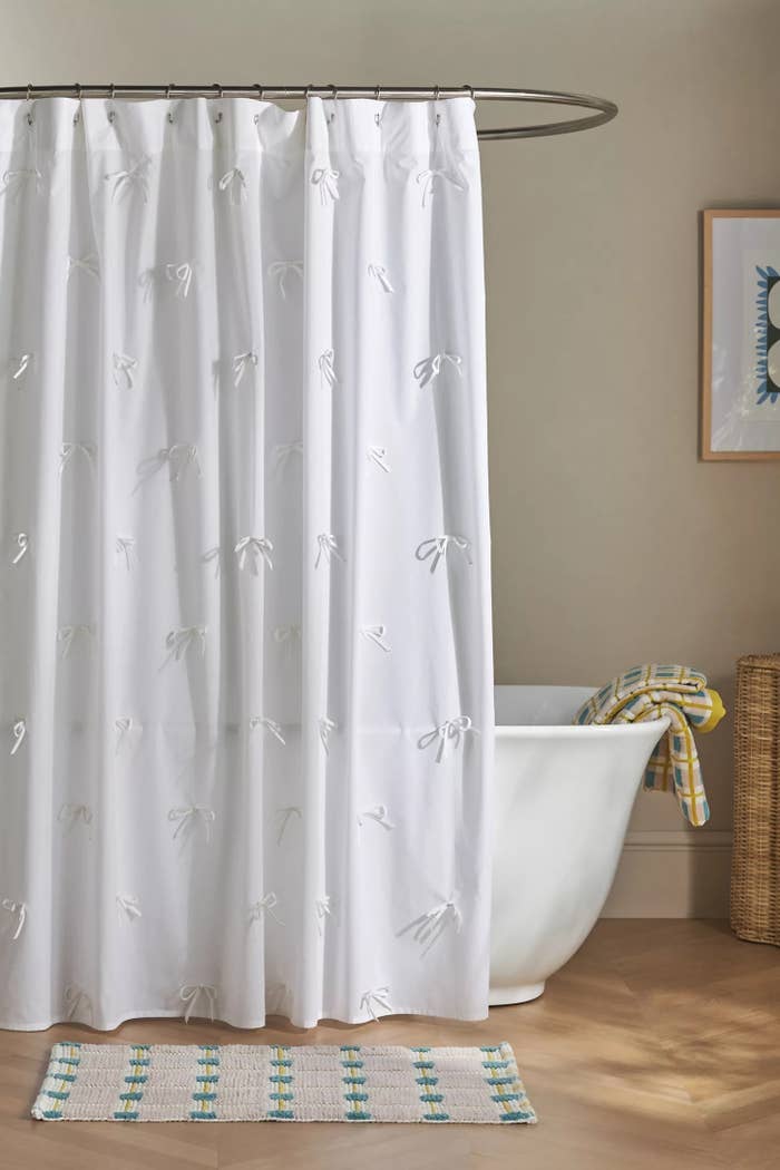 White shower curtain with bow patterns hangs in a bathroom with a freestanding tub, wicker basket, and patterned floor mat