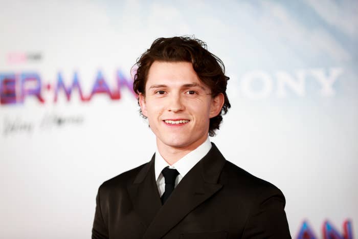 Tom Holland in a formal suit smiles on a red carpet at a movie premiere event