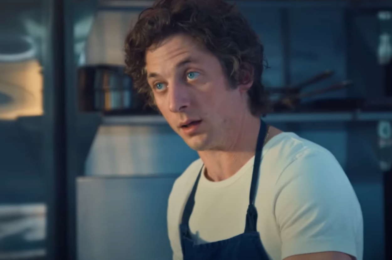 Person wearing a white t-shirt and dark apron standing in a kitchen, appearing focused and engaged