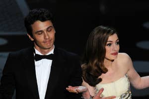 Two people on stage shrugging. James Franco wears a tuxedo; Anne Hathaway is in an embellished white gown