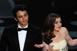 Two people on stage shrugging. James Franco wears a tuxedo; Anne Hathaway is in an embellished white gown