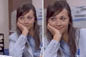 A person in a suit rests their chin on their hand, smiling slightly while sitting at an office desk