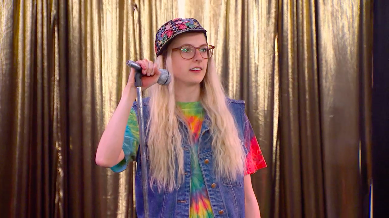 Person on stage wearing a tie-dye shirt, jean vest, floral cap, and glasses, holding a microphone