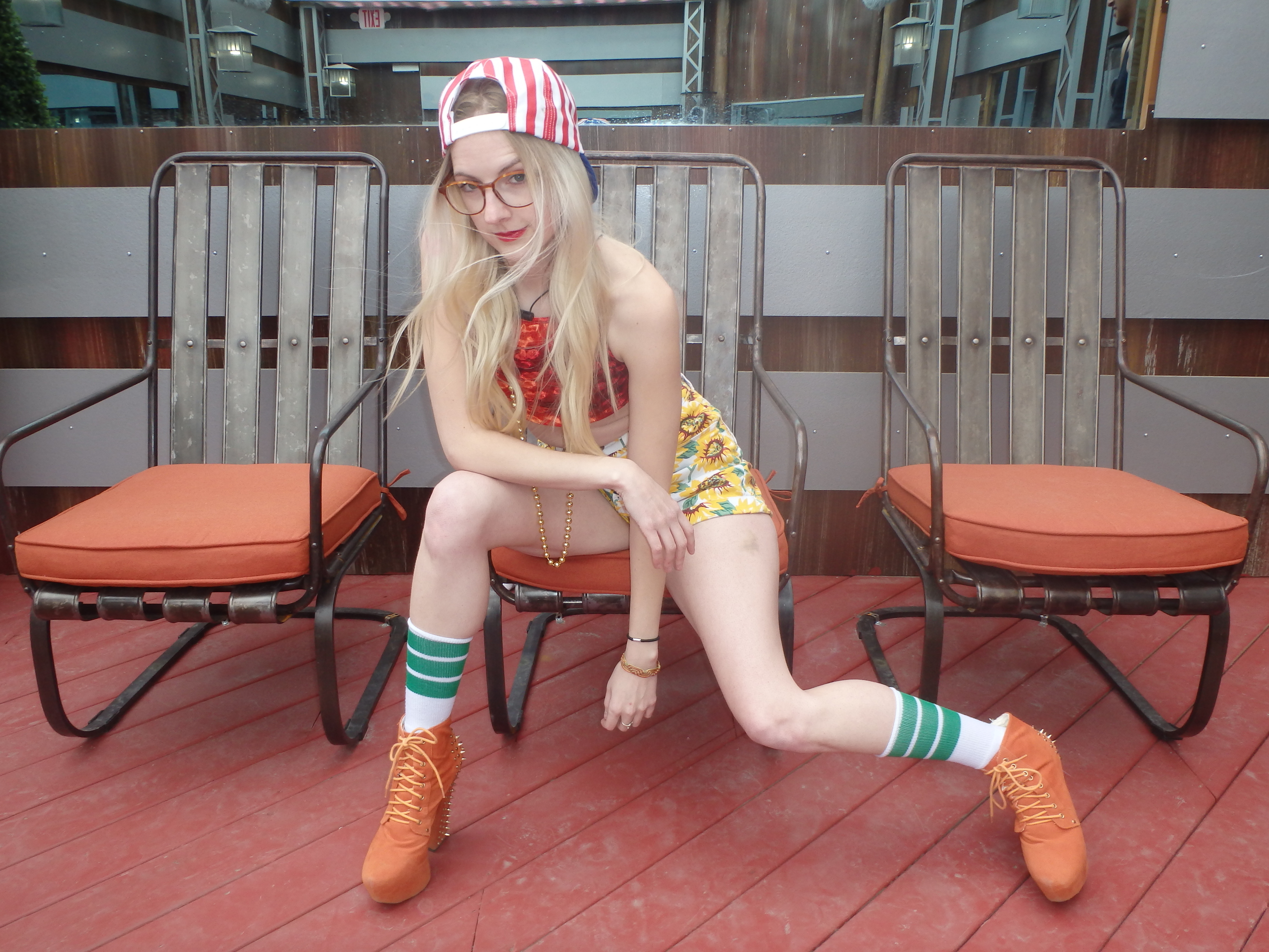 Person in a playful pose on a deck wearing a patterned outfit, striped socks, and red boots, with a striped cap turned backward