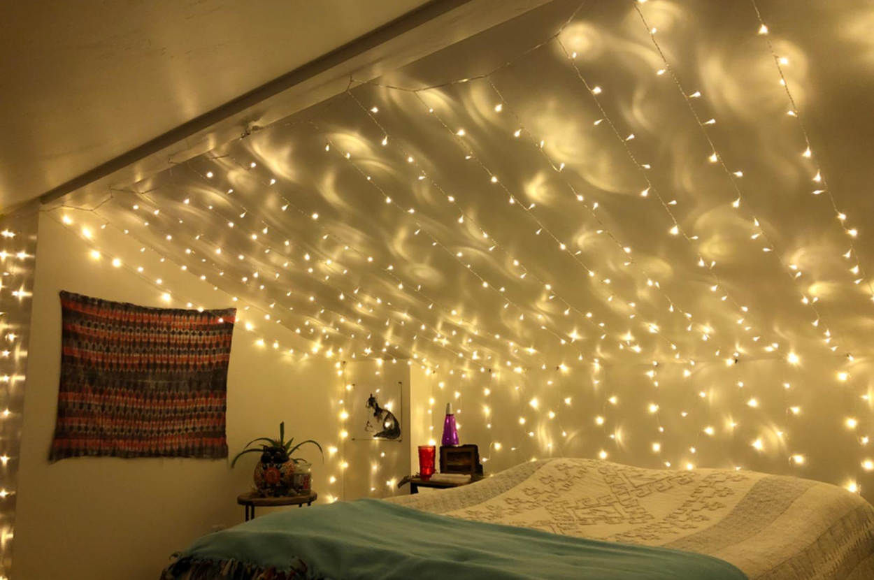 Cozy attic bedroom with string lights on the ceiling, a woven wall hanging, a bed with layered blankets, and a small nightstand with decor