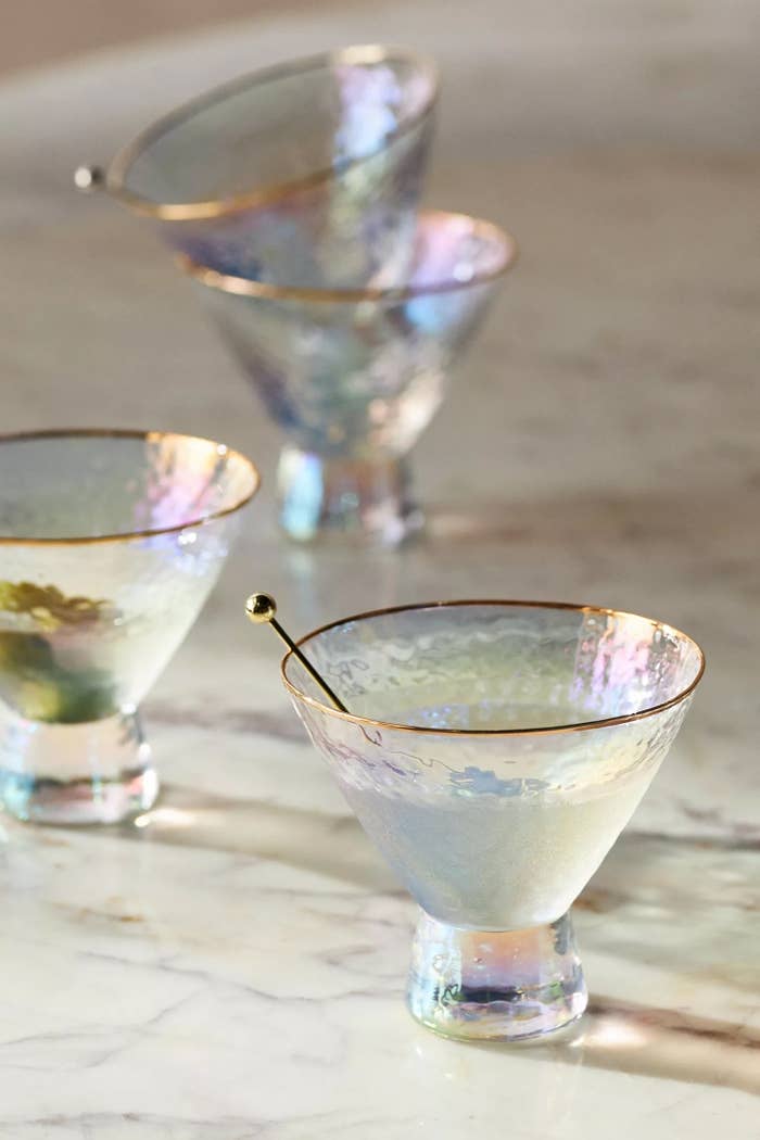 Four iridescent martini glasses with gold rims and cocktail picks rest on a marble surface. Ideal for stylish home entertaining