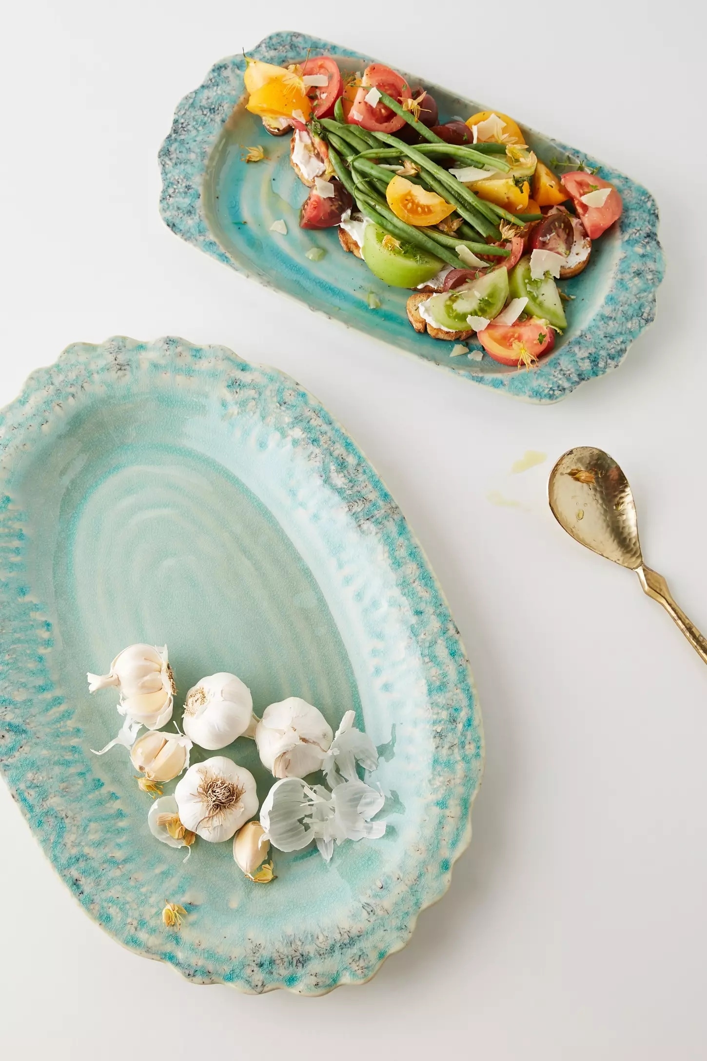 Two oval ceramic platters: one with a fresh vegetable salad and the other holding garlic bulbs. A gold spoon lies beside them