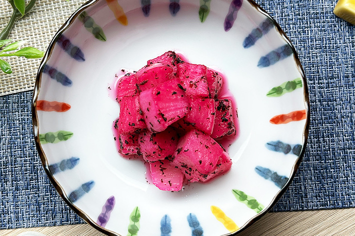 縁に模様のある皿に盛られたピンクの漬物。箸と植物が横に配されている。