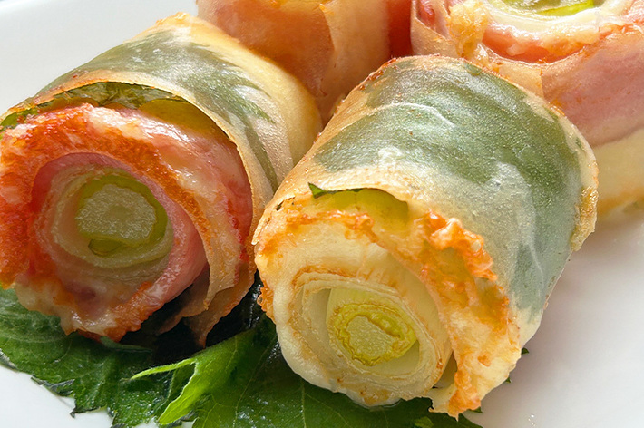 ネギとベーコンを巻いた春巻きが白い皿に盛られています。葉も添えられています。