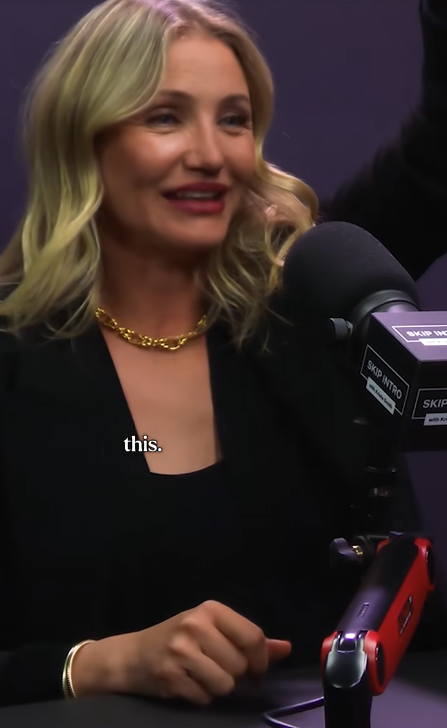 Cameron Diaz speaking into a microphone during an interview, wearing a black outfit with a gold necklace