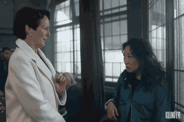 Sandra Oh in Killing Eve makes a face and waves goodbye to a woman who stands looking shocked in a white jacket