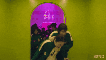 Contestants in green tracksuits run through a vibrant room with pink lighting, looking nervous as they hide and close the door in Squid Game