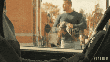 A scene from the series &quot;Reacher&quot; showing a muscular man in a tight shirt wrapping his hand in a fabric before punching through a driver side car window