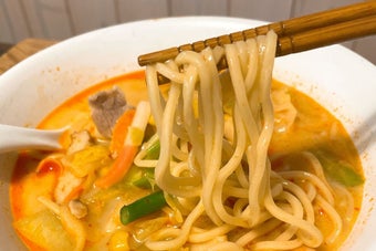 箸でつまんだラーメンの麺が白い丼に盛り付けられ、野菜と肉が入ったスープが見える。