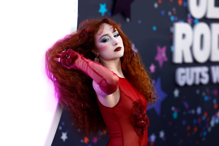 Chappell Roan with long red hair, wearing a sheer, red floral gown. They are posing dramatically at a celebrity event