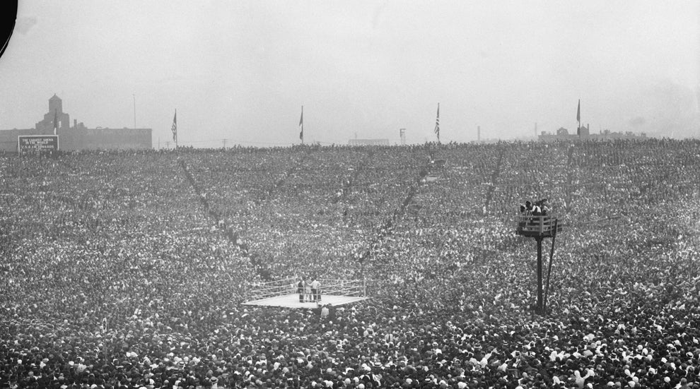 Aerial view of a crowded stadium centered on a boxing ring, filled with spectators, creating a sea of hats in a historical black-and-white photograph