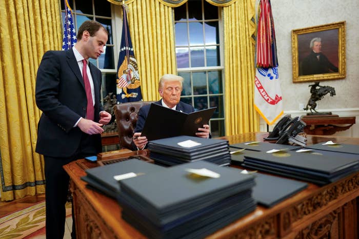 A person sits at a desk filled with folders, reviewing documents, while another person stands nearby in an office setting