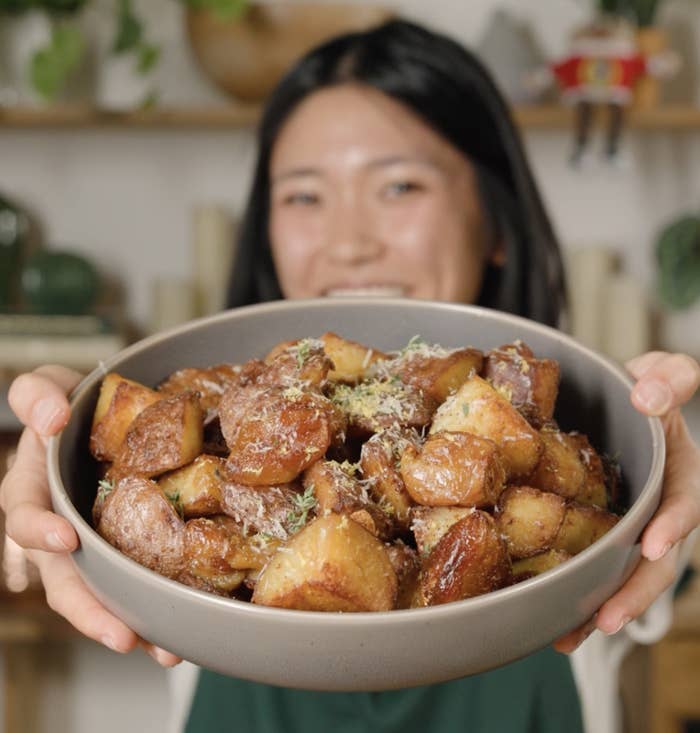Person holding a vessel  of crispy roasted potatoes garnished with herbs and grated cheese, smiling successful  a cozy room  setting