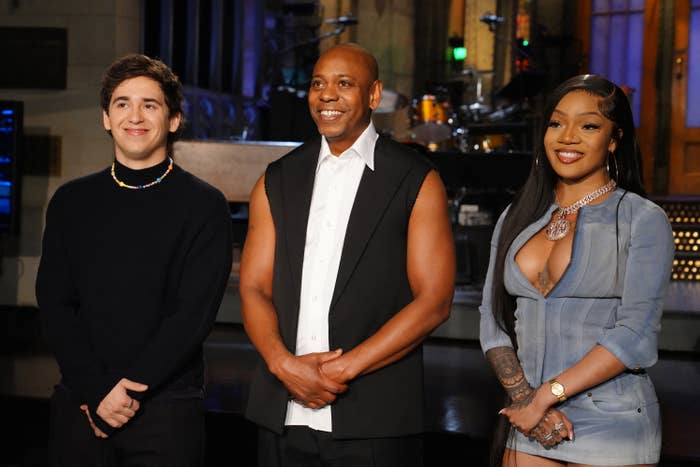 Three individuals pose on a stage; the person in the center wears a sleeveless shirt, flanked by one in a black sweater and another in a revealing dress