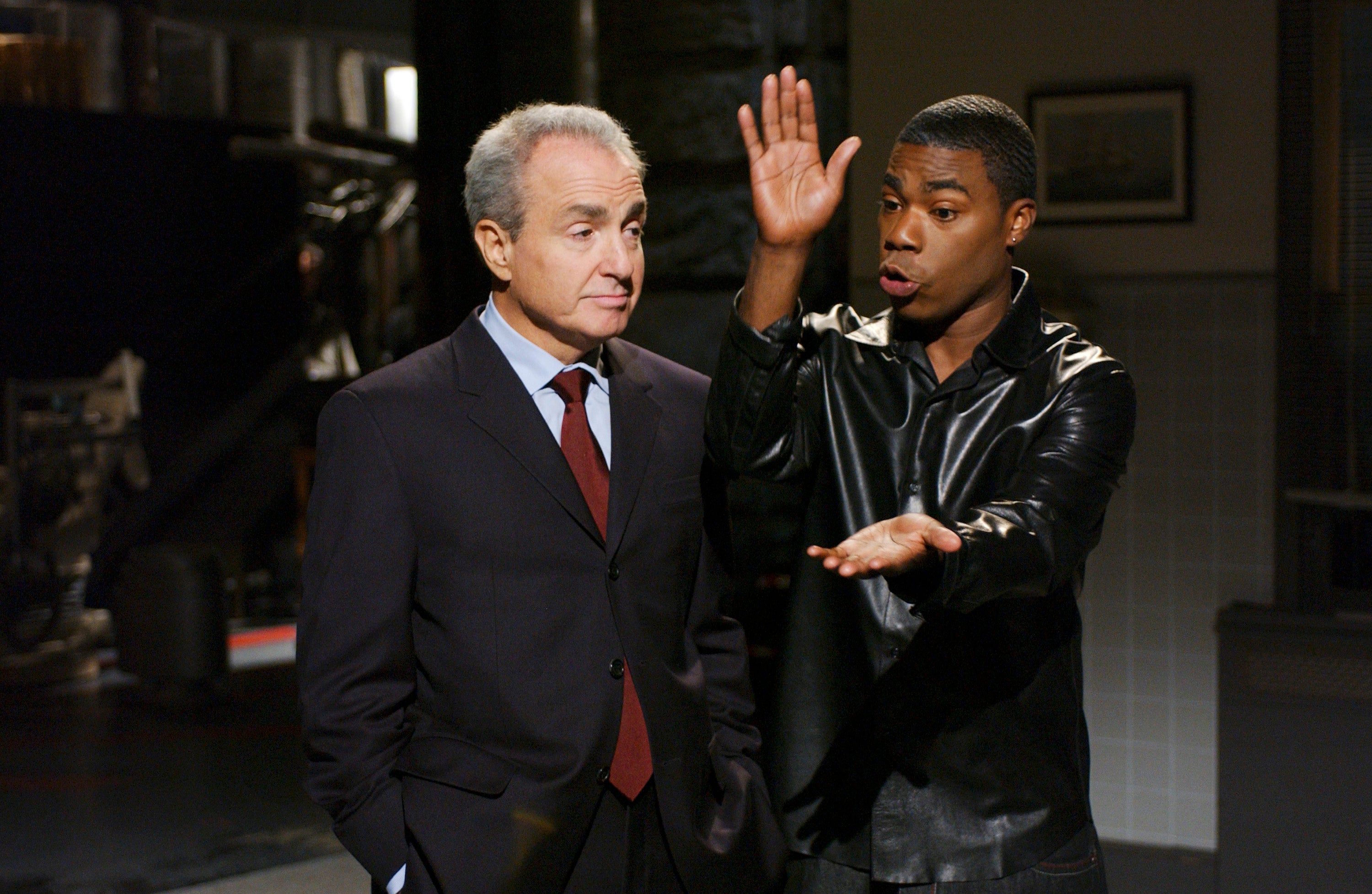 Two people are engaged in a discussion on a set. One gestures animatedly while the other listens attentively in a suit