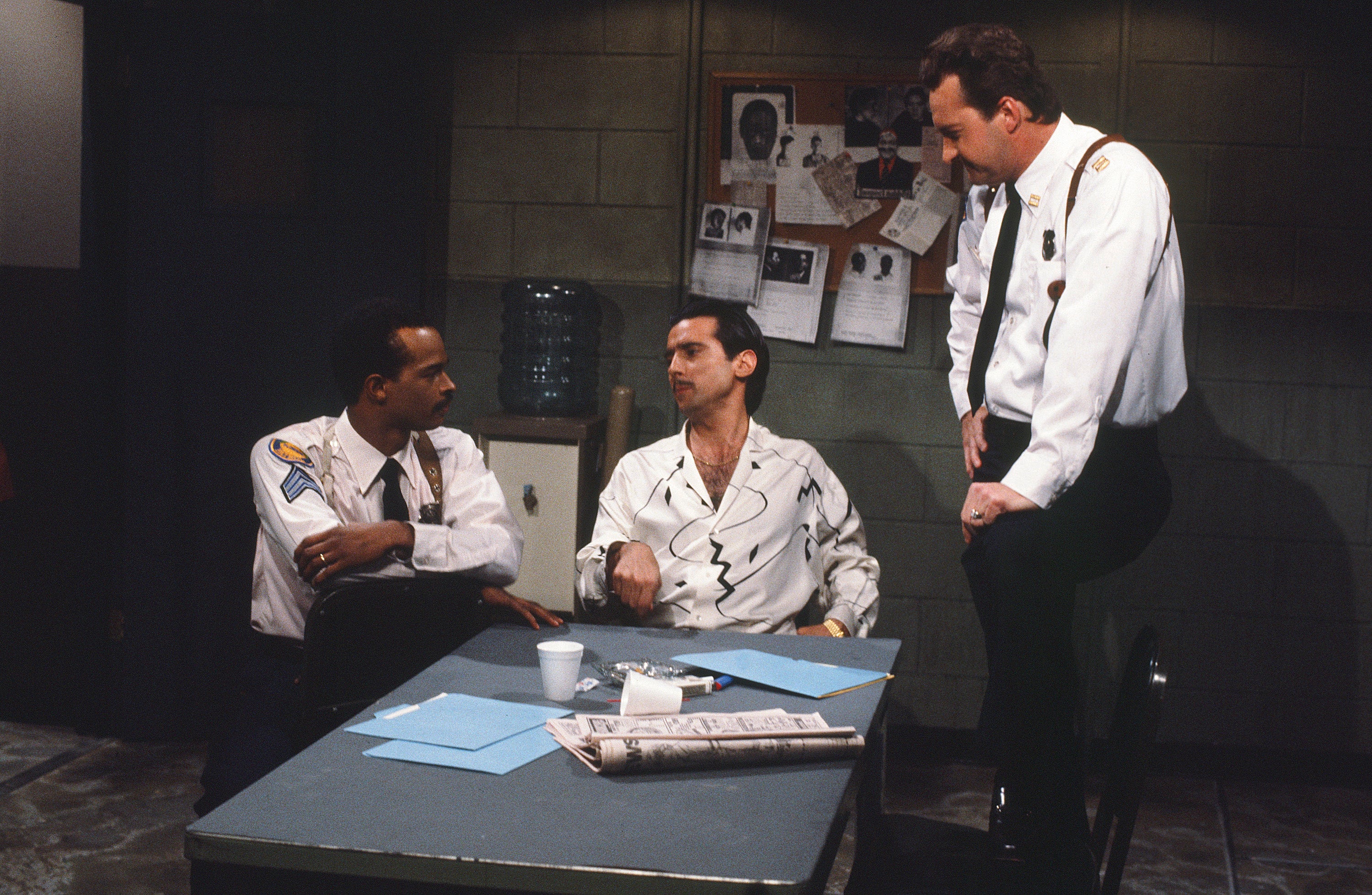 Three men in police uniforms are engaged in a conversation around a table in a police station setting