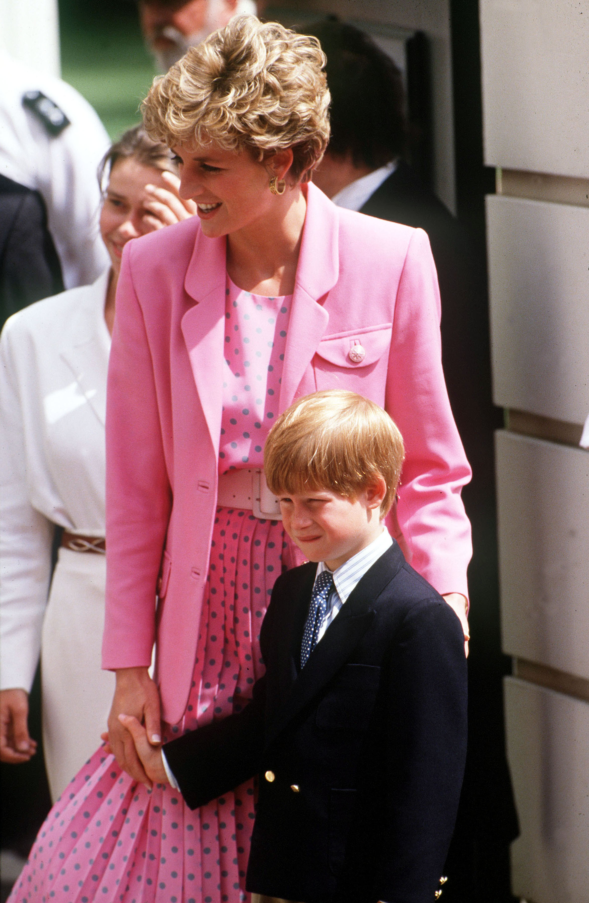 Princess Diana in a polka dot dress and matching blazer holding hands with Prince Harry in a navy blazer and tie, both smiling outdoors