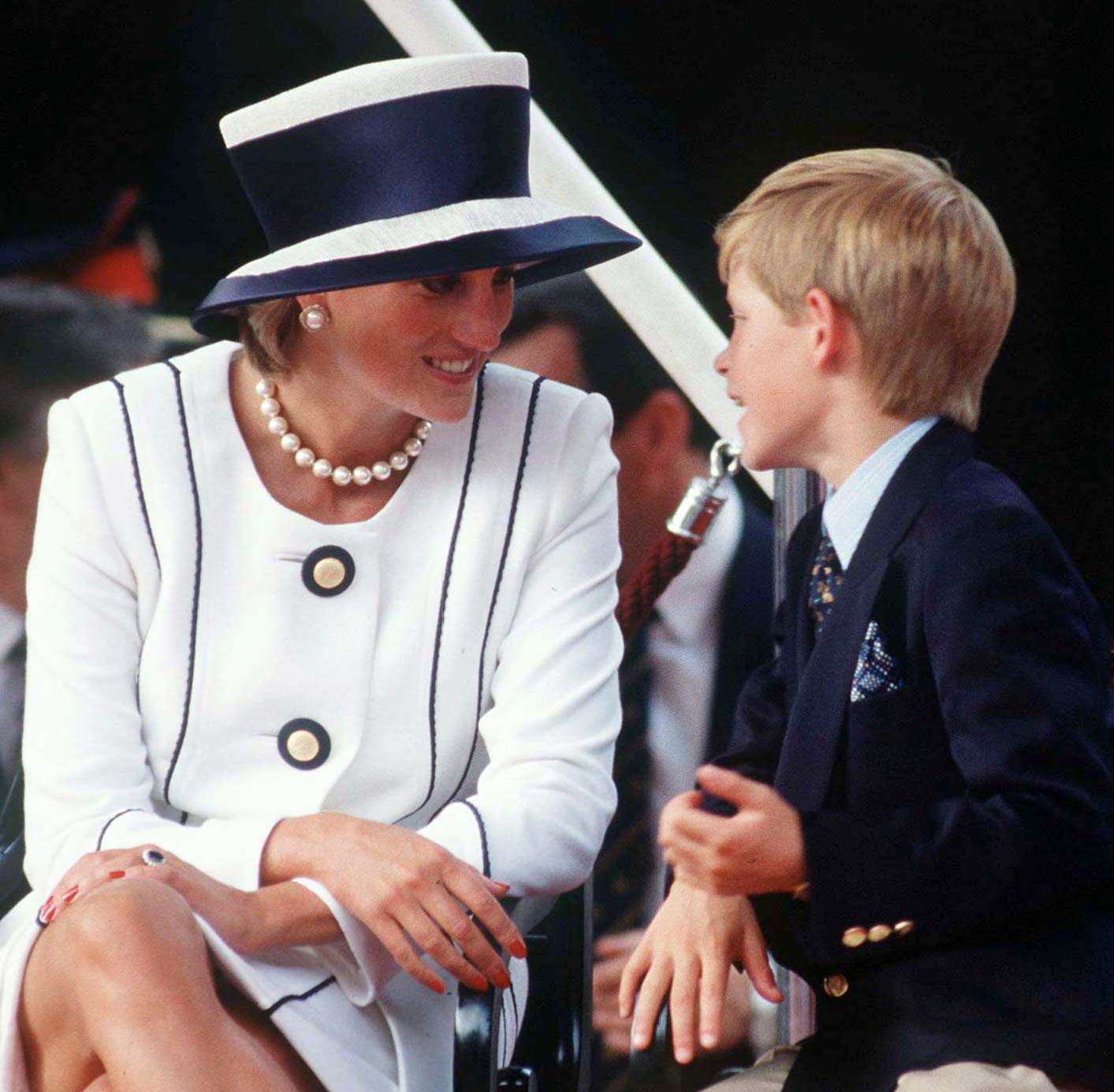 Princess Diana in a stylish white outfit with a wide-brimmed hat smiles at Prince Harry in a suit as they converse