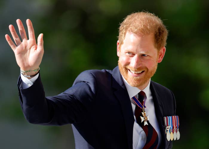 Prince Harry in formal attire waving, wearing a suit with various medals and a tie