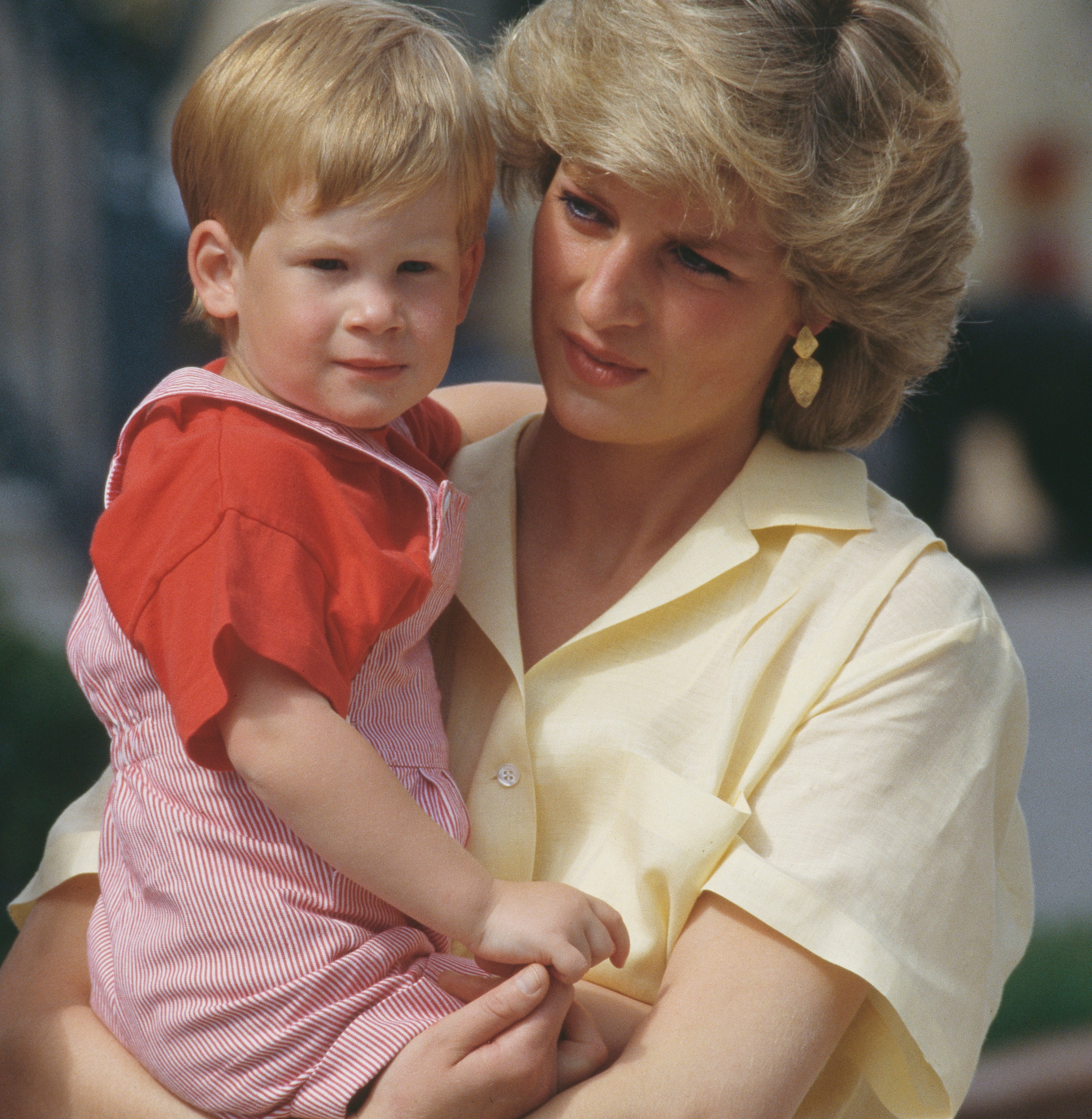 Princess Diana holds young Prince Harry in her arms outdoors. She is wearing a short-sleeve blouse, and the child is in a striped outfit