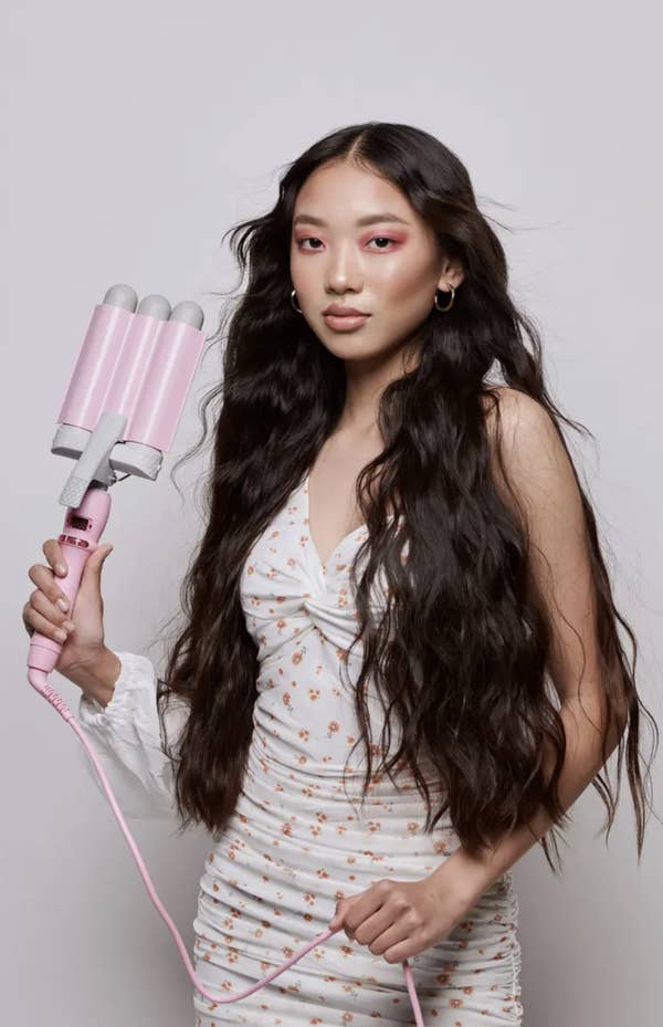 Person with long wavy hair using a triple barrel hair waver. They&#x27;re wearing a white floral dress with ruffled details