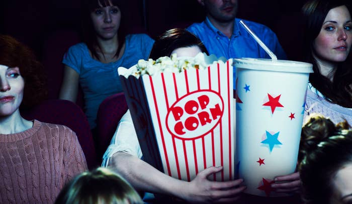 Person in a cinema holds oversized popcorn and drink, obscuring their face, surrounded by other moviegoers