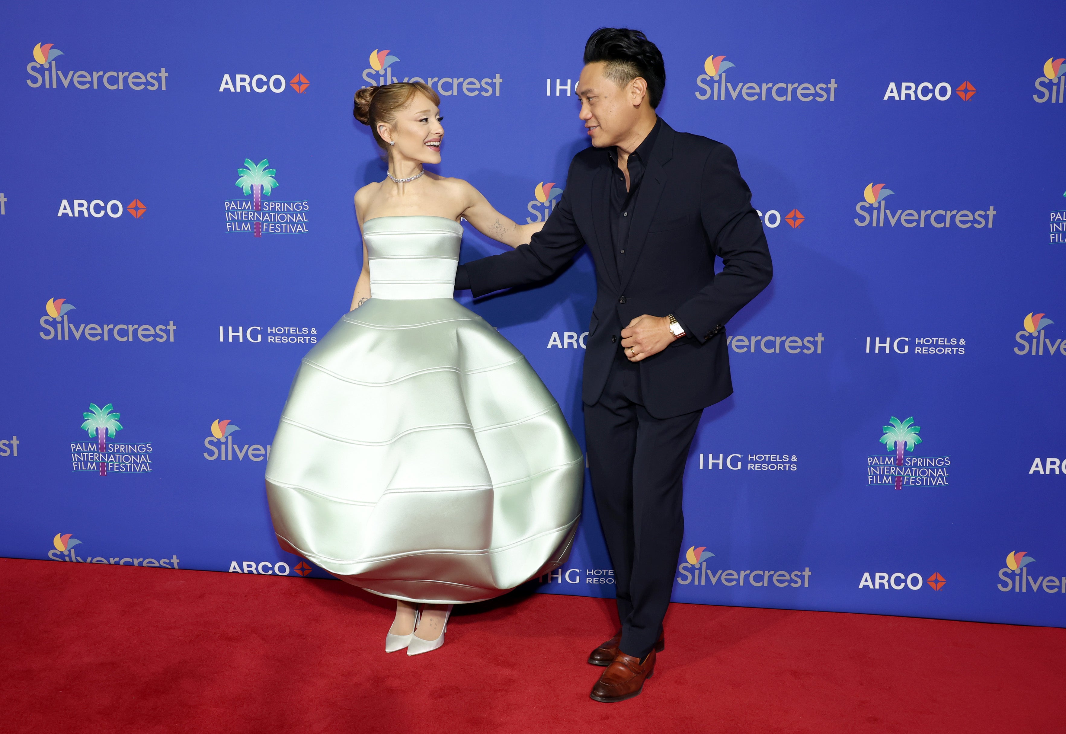 Two individuals stand on a red carpet. The woman is in a structured metallic ball gown, and the man is wearing a classic suit