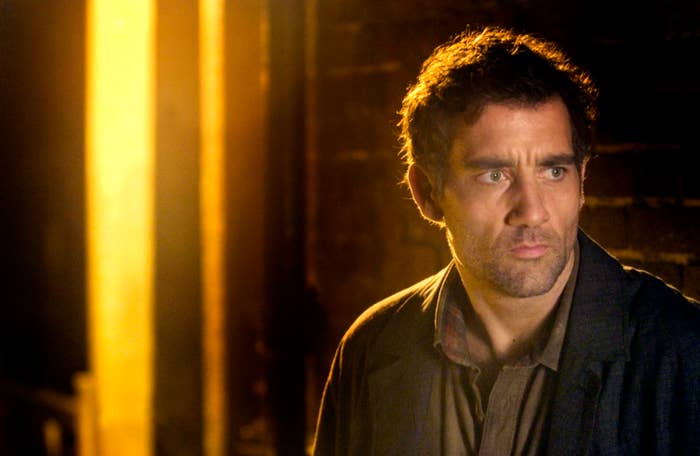 A contemplative man stands by a brick wall, partially lit by a bright light, wearing a dark jacket and shirt