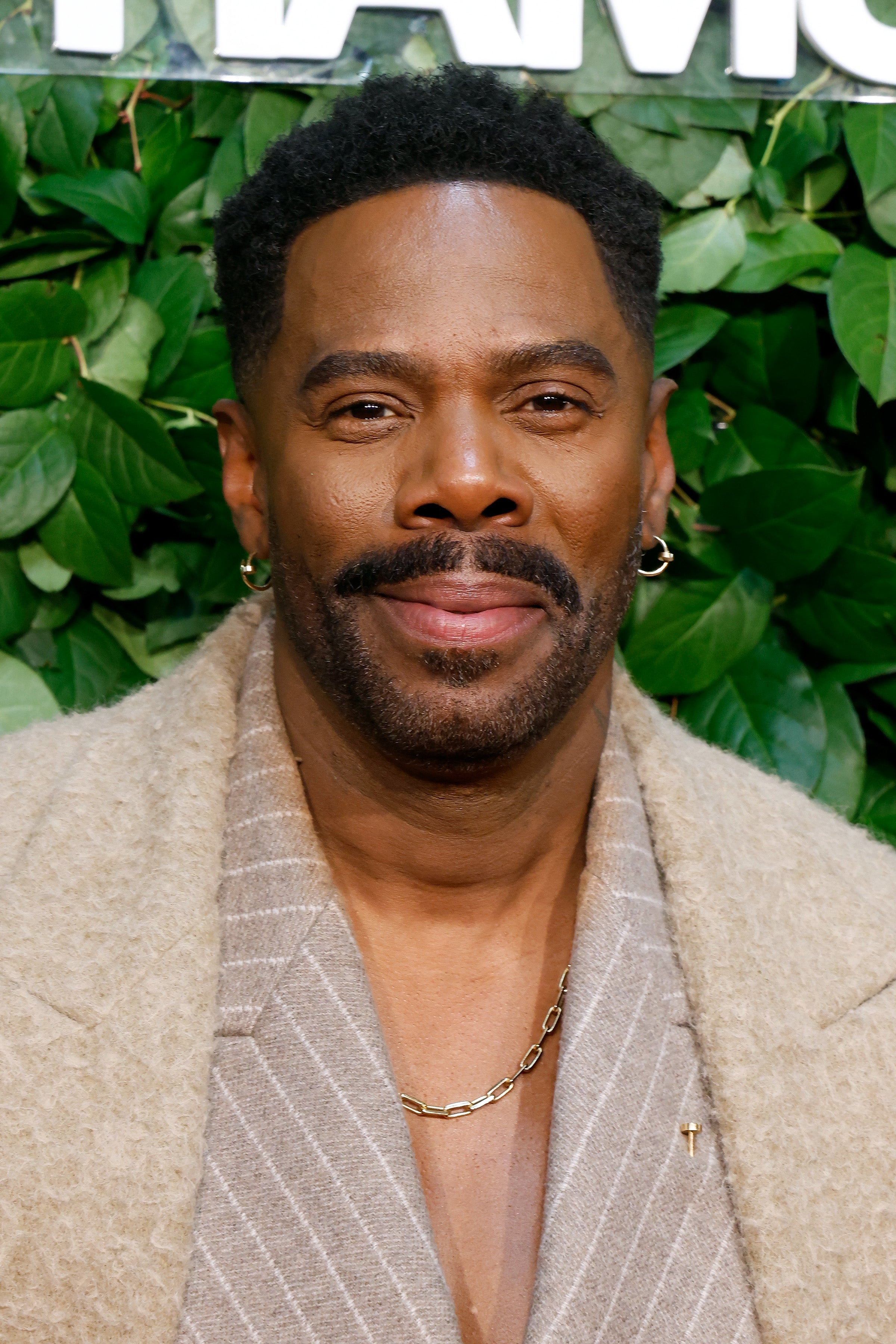 Colman Domingo wearing a textured coat, striped shirt underneath, and gold chain, posing in front of leafy backdrop