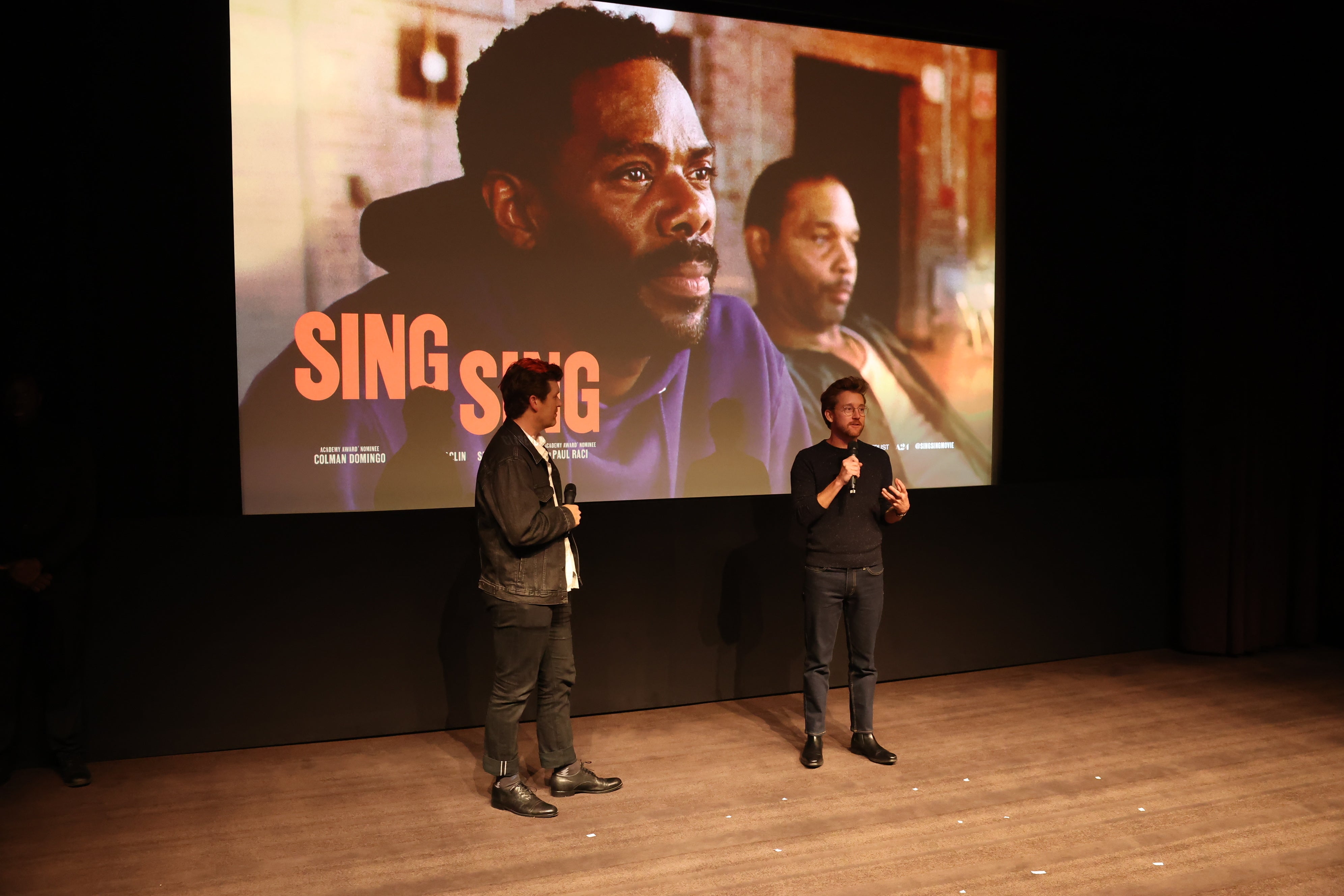 Two people stand on stage speaking, with a large screen behind showing a film scene from "Sing Sing."