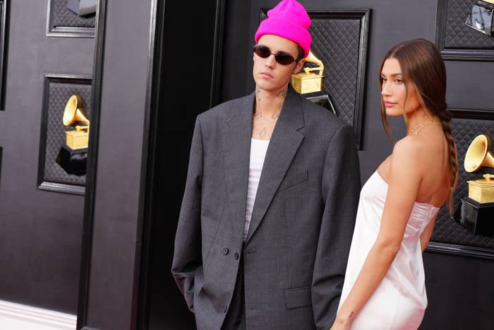 Justin Bieber in an oversized suit and beanie stands with Hailey Bieber in a strapless gown on a red carpet, posing near stylized award displays