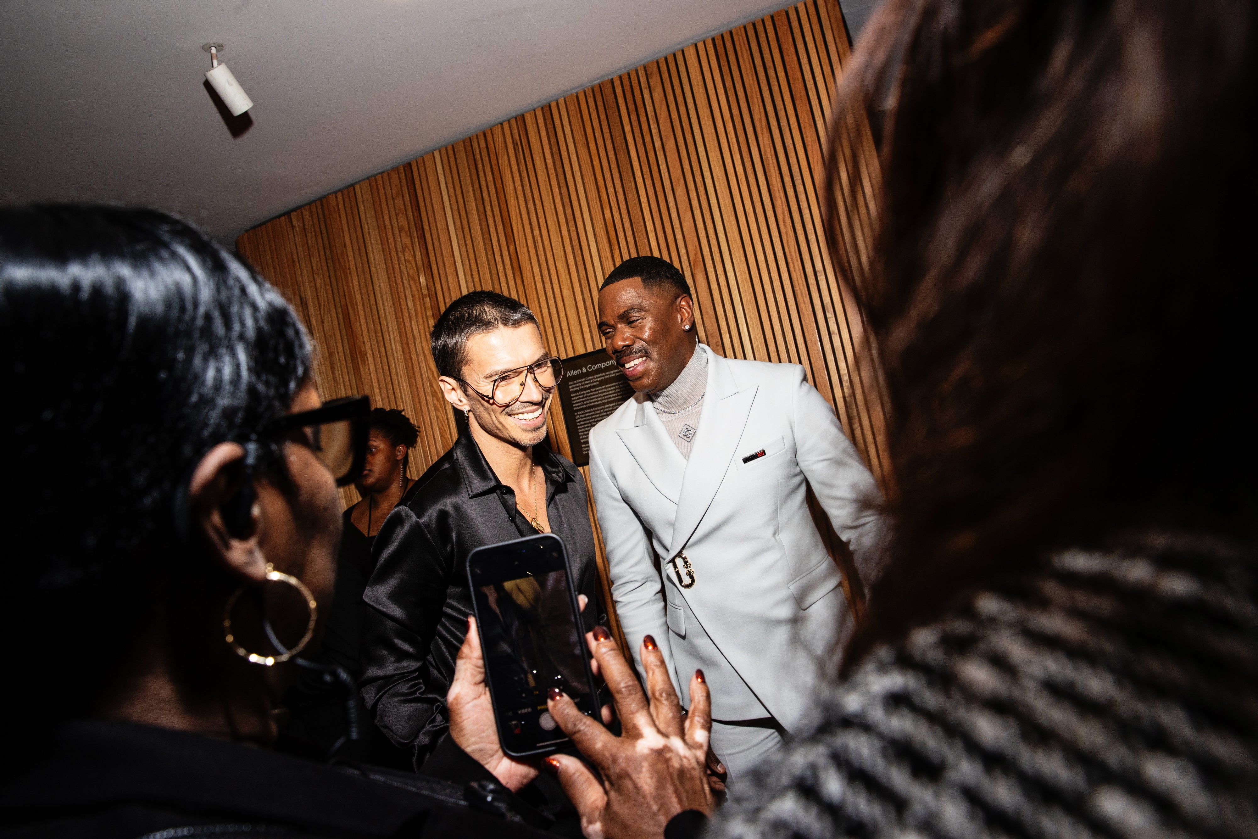 Colman and Raúl smiling and talking at an event, with one wearing a dark outfit and the other in a light suit. Attendees are capturing the moment
