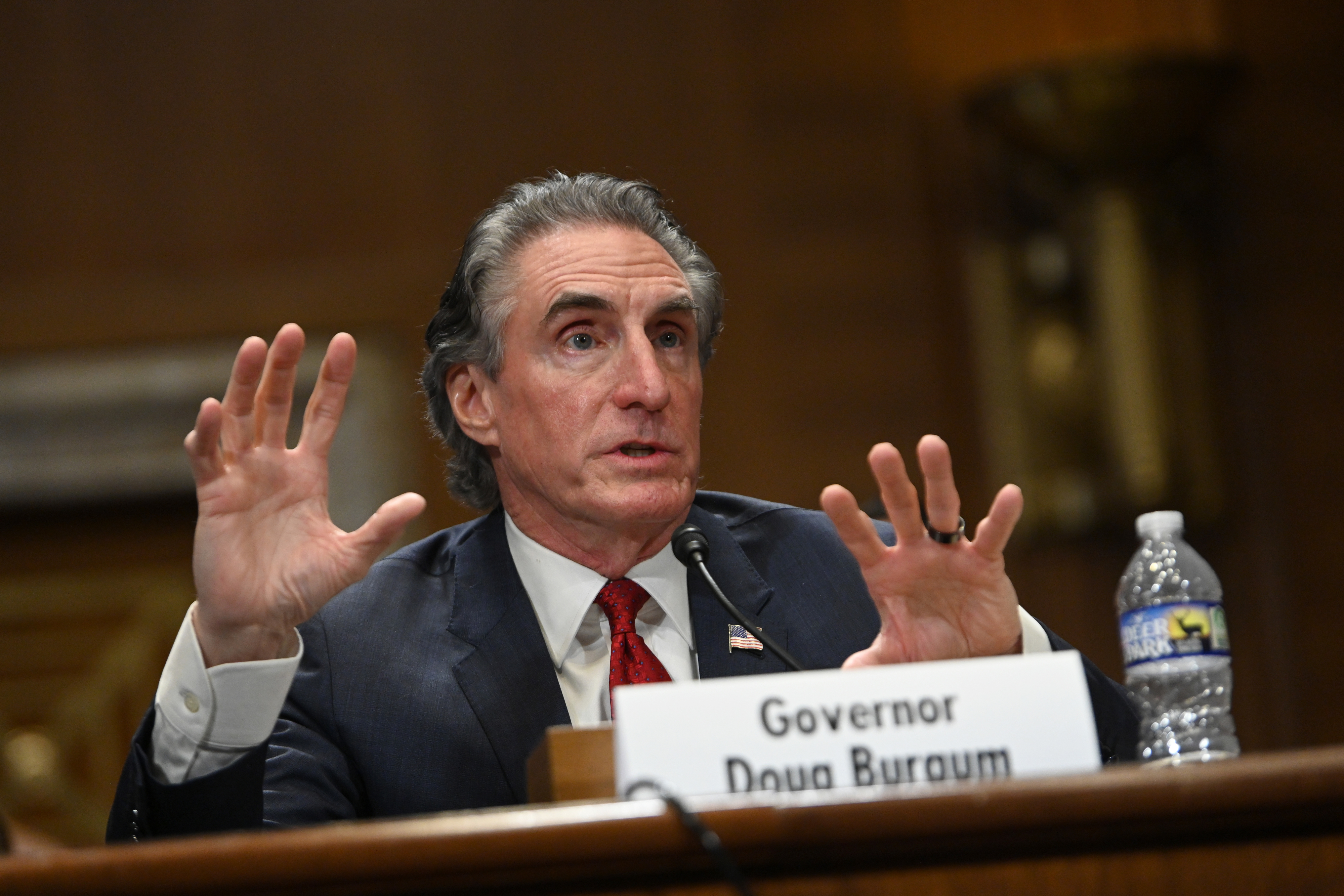 Man in a suit speaking and gesturing with his hands at a conference or meeting, seated behind a nameplate reading "Governor Doug Burgum."